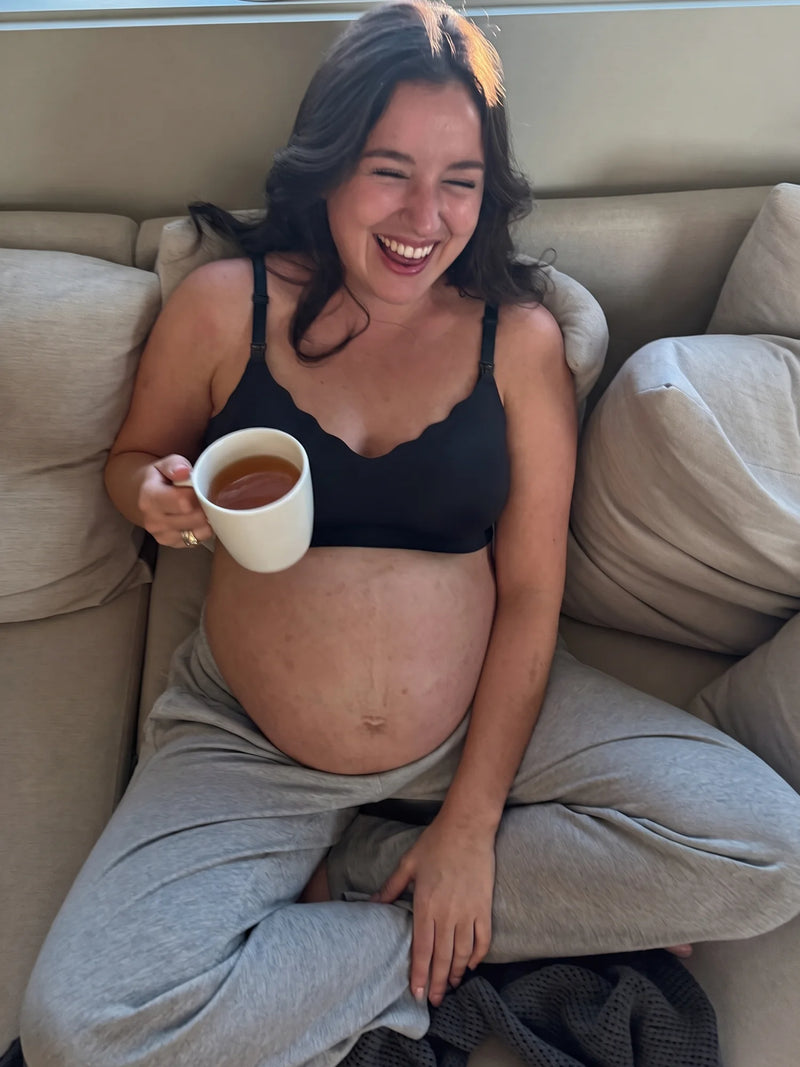 Pregnant mom laughing on the sofa with tea in the Aura Comfort Bra in Black